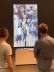 Zwei Schüler stehen vor einem großen Bildschirm. Der Bildschirm zeigt wie sie aussehen würden wenn sie gezeichnet werden
